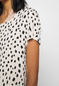 Top a maniche corte in tessuto beige chiaro con un motivo astratto a pois neri. Presenta un collo rotondo e una vestibilità comoda. Tessuto morbido.