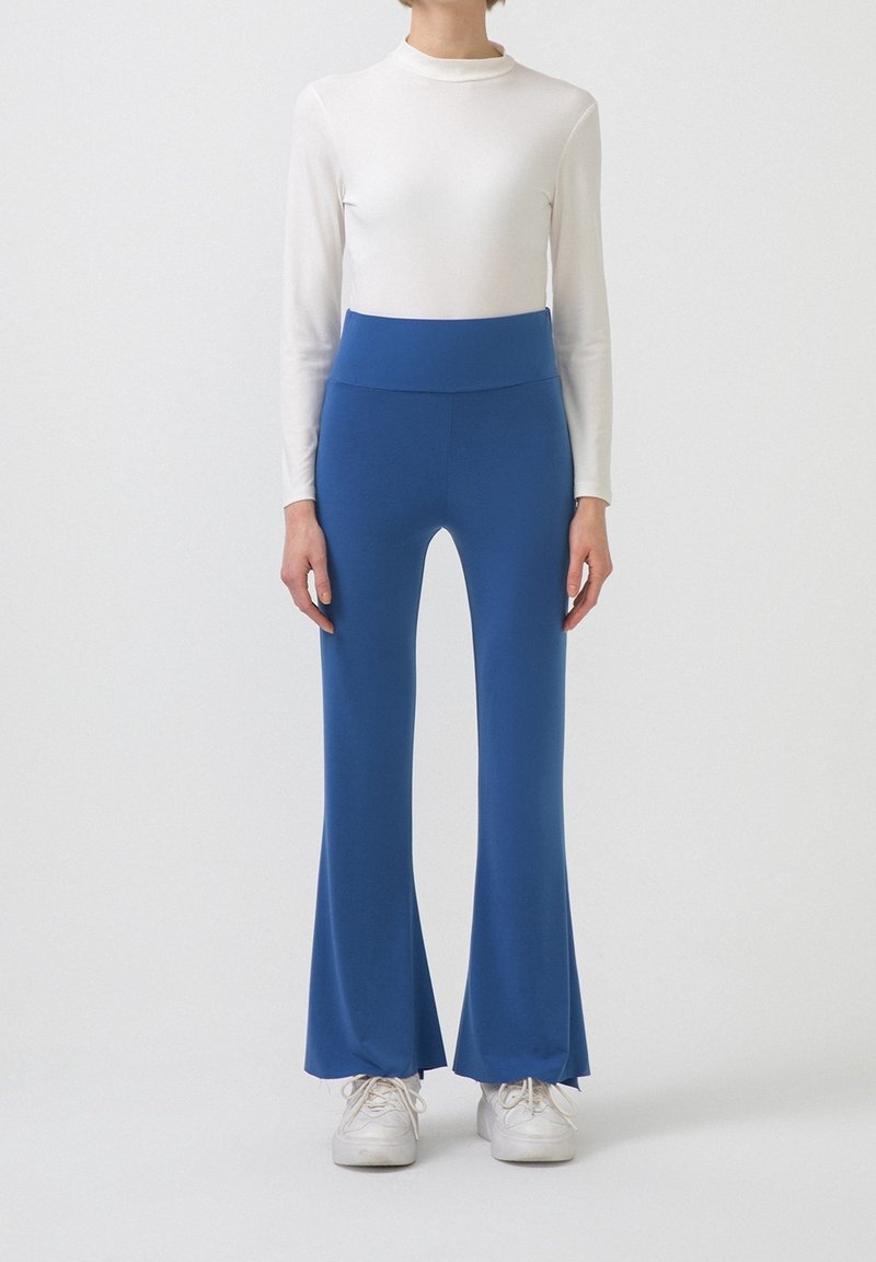 Femme portant un pantalon évasé bleu taille haute, un haut à manches longues blanc et des baskets blanches, se tenant devant un fond blanc uni.