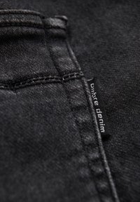 Tissu en denim noir avec un gros plan sur une couture et une étiquette portant l'inscription « ombre denim ». La texture semble légèrement usée, avec des variations subtiles de teinte.