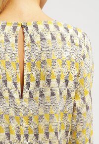 Blusa amarilla y gris con un estampado, fabricada con un tejido texturizado, que presenta un detalle de escote en forma de lágrima en la espalda y un cierre de botón en el cuello.
