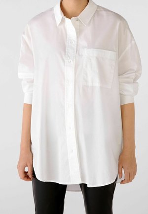 Persona che indossa una camicia bianca ampia con taschino sul petto e maniche arrotolate, abbinata a pantaloni neri.