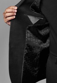 Schwarze Anzugjacke mit strukturiertem Äußeren, ausgestattet mit einem eleganten Satinfutter und Taschen-Details. Das Innere zeigt ein geometrisches Muster.