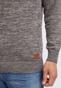Brun strikket genser med et teksturert mønster, ribbede mansjetter og kant, samt en brun skinnlabel nederst til venstre.
