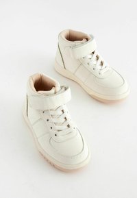 Baskets montantes blanches en cuir lisse avec une sangle texturée, des lacets et des semelles beige clair. Design simple avec de légers détails de couture.