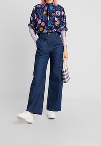 Blouse à motif floral avec manches courtes, associée à un jean en denim foncé taille haute à jambes larges. Le modèle porte des baskets blanches et tient un sac à carreaux.