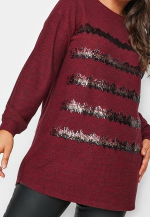 Woman wearing a burgundy long-sleeve sweater with five horizontal black and silver sequin stripes, paired with black pants.