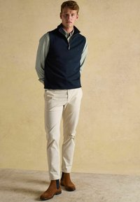 Námornícky v krku bez rukávov nad svetlozelenou pruhovanou košeľou, doplnený krémovými nohavicami a hnedými semišovými Chelsea čižmami.