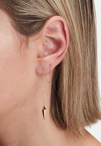 Gol Symbole mit einem Kettendesign, das einen spitzen geometrischen Anhänger und einen geraden Stab umfasst und von einem Ohrläppchen in Kurzen Haaren hängt.