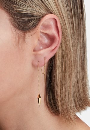 Boucles d'oreilles en or présentant un design en chaîne avec un pendentif géométrique pointu et une barre droite, suspendues à un lobe d'oreille contre des cheveux courts.