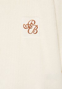 Tissu tricoté couleur crème avec un petit patch brodé présentant les lettres cursives entrelacées "J" et "B" en fil marron.