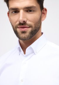 Witte button-up shirt met een spread collar, voorzien van kleine witte knopen en een gladde textuur. Model toont een schone, neutrale uitdrukking.