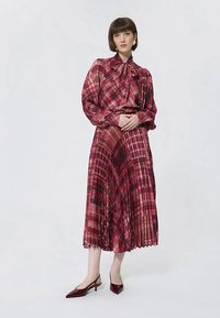 Mulher em pé com uma blusa de manga longa em padrão de xadrez vermelho e rosa, com uma gravata, e uma saia midi plissada a condizer, usando sapatos de salto baixo vermelho escuro.