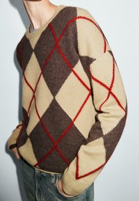 Pull en tricot avec un motif de losanges en marron, crème et rouge, avec un col rond côtelé et une coupe décontractée.