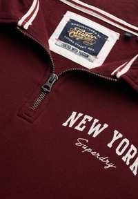 Bordowa bluza rozpinana z białym kołnierzem w czerwone paski, z przednim zamkiem błyskawicznym oraz nadrukowanym napisem "NEW YORK" i "Superdry" w białym kolorze.
