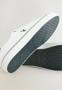 Witte sneakers met een gestructureerde zwarte rubberen zool. Het bovenwerk heeft een gladde stof en subtiele logo-details. Veters en minimalistisch ontwerp.
