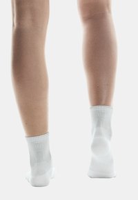 Weiße Knöchelsocken mit gerippter Textur und verstärktem Fersenbereich. Die Ansicht zeigt die Rückseite der Socken an Beinen vor einem hellen Hintergrund.