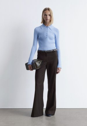 Hellblaues geripptes Langarmshirt mit Kragen, braune weite Hose und eine dunkel gefärbte Clutch-Tasche mit strukturierten Akzenten.