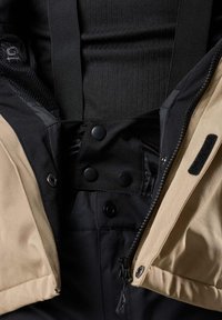 Schwarze und beige Oberbekleidung mit verstellbaren Trägern, Druckknöpfen und Reißverschlüssen. Strukturiertes Innenfutter mit glattem Stoff und Klettverschluss-Elementen.