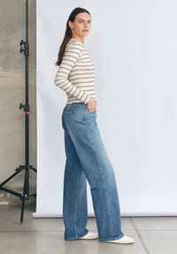 Striped top z długim rękawem w kolorze kremowym i granatowym, w połączeniu z jasnoniebieskimi spodniami typu wide-leg. Modelka stoi bokiem, ma na sobie białe buty. W tle znajduje się regulowane tło.