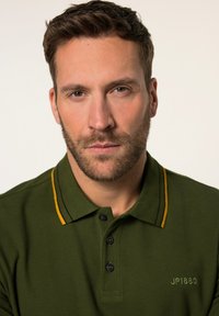 Tmavě zelené polo triko s žlutým akcentem na límci, s třemi knoflíky a vyšitým nápisem "JP1880" v zelené barvě.