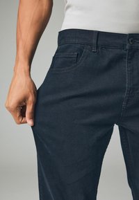 Donkerblauwe denim broeken met een slim fit, voorzien van een zijzak en een knoopsluiting. Het materiaal lijkt glad met subtiele stikseldetails.