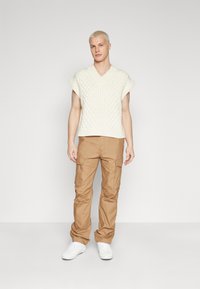 Strikkegenser i hvitt med V-utringning og korte ermer, sammen med beige cargo-bukser. Hvite sneakers fullfører antrekket.