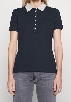 Femme portant un polo bleu marine ajusté avec un col à carreaux blancs et des boutons blancs, associé à un jean gris.