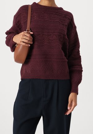 Personne portant un pull en laine texturé bordeaux foncé, un pantalon noir, et tenant un sac à bandoulière marron sur l’épaule.