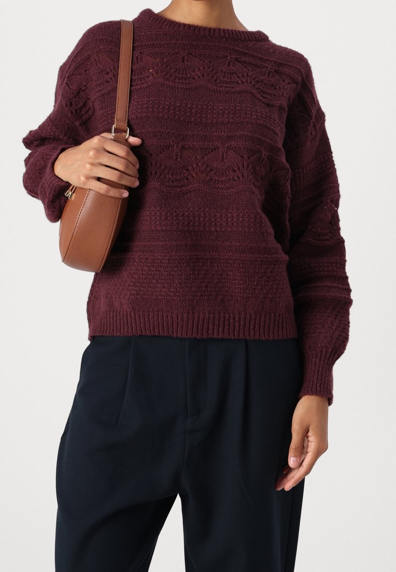 Personne portant un pull en laine texturé bordeaux foncé, un pantalon noir, et tenant un sac à bandoulière marron sur l’épaule.