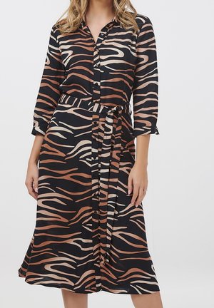 Kobieta ubrana w sukienkę koszulową w czarno-brązowo-beżowe paski zebry, z rękawami do połowy przedramienia i pasem z tego samego materiału w talii.