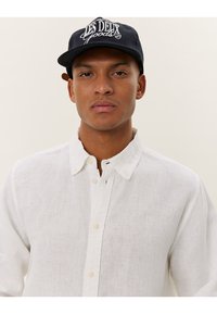 Camicia bianca in lino con colletto, caratterizzata da un leggero logo in rilievo. Abbinata a un cappello blu navy con testo bianco.