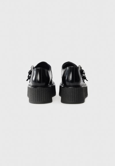 Black leather platform shoes viewed from the back with thick textured soles and silver buckle details on a plain background.