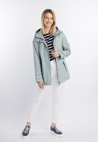 Ljusgrå vattentät jacka med huva, dragkedja och tryckknappar. Randig skjorta under, ihop med vita byxor och mörka sneakers.