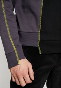 Sudadera gris y negra con costuras en neón verde contrastante a lo largo de las costuras, con una tela texturizada, mangas acampanadas y un diseño ajustado.