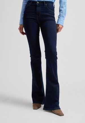 Persona, kas valkā tumši zilas izplūdušas džinsas, gaiši zilu kreklu un brūnas kurpes ar smailiem purngaliem, stāv uz gaiši pelēka fona.