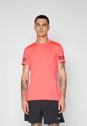 T-shirt de sport corail à manches courtes avec un col rond et logo sur la manche gauche ; le tissu semble lisse et ajusté.