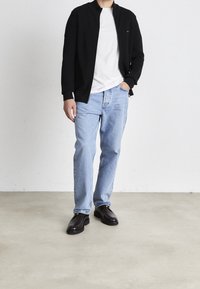 Man står klädd i svart zip-jacka, vit skjorta, ljusblå jeans och mörkbruna läderskor på en enkel golvyta med vit vägg i bakgrunden.