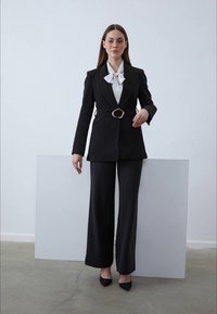 Svart skräddarsydd kostym med vida byxor och en knäppt blazer. Vit blus med fluga under, kompletterad med svarta klackar. Enfärgad bakgrund.
