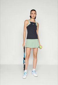 Joven mujer con un conjunto de tenis negro y menta, sosteniendo una raqueta y una pelota, de pie sobre un fondo gris claro liso.