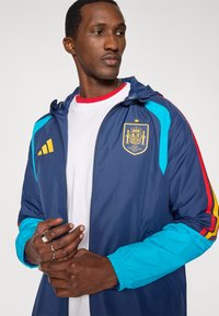 Marineblauwe windjack met blauwe en rode accenten, voorzien van een gouden Spaans wapenschild. Het materiaal is lichtgewicht en heeft een verstelbare capuchon.