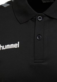 Sort polo-shirt med tekstureret stof, tre-knaps lukning og et hvidt logo på venstre side. Inkluderer kontrasterende geometrisk detalje på skuldrene.