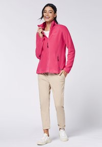 Fleece jacket in bright pink with high collar, full zip, and two zippered pockets. Paired with beige pants and white sneakers.