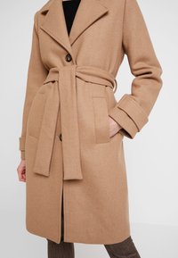 Manteau ceinturé en mélange de laine camel, doté d'un col à revers, d'une fermeture à bouton unique et de deux poches frontales, avec une texture lisse.