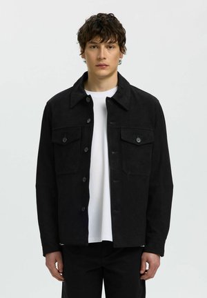 Jonge man met kort krullend haar draagt een zwarte jas met knopen over een wit shirt, staand tegen een effen lichte achtergrond.
