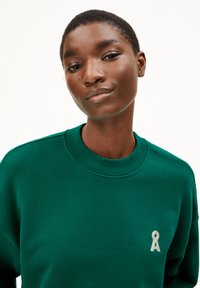 Grüner Sweatshirt mit Rundhalsausschnitt, Textilstruktur und einem kleinen hellgrauen Emblem auf der Brust. Schlichtes Design, lässiger Stil.