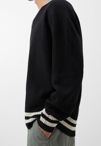 Czarny sweter z dzianiny z dekoltem w serek, z kontrastującymi białymi i beżowymi paskami na mankietach i dole. Luźny krój.