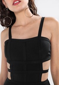Svart kort topp med breda axelremmar, strukturerade paneler och utskärningar på sidorna. Tyget verkar texturerat med en figurnära design.