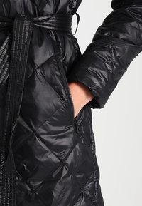 Svart puffjacka med diamant-quilting, långa ärmar och midjebälte. Har en sidodragkedjeficka och glänsande materialtextur.