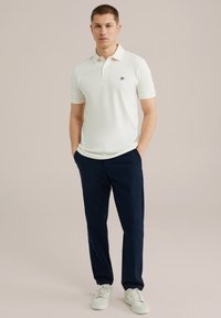 Bílá polo košile s krátkým rukávem, námořnické kalhoty a bílé tenisky. Polo má jemné logo na hrudi. Hladká textilie.