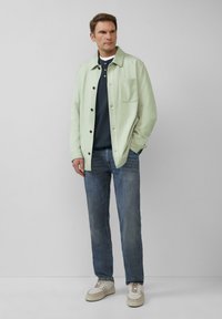 Lichtgroene jas met een knoopsluiting aan de voorkant en een borstzak, een marineblauwe shirt eronder, blauwe spijkerbroek en witte/creme sneakers.